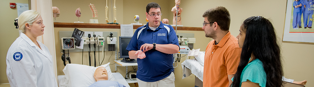 A professor at Penn State Worthington Scranton talks to two nursing students in a classroom hospital room.